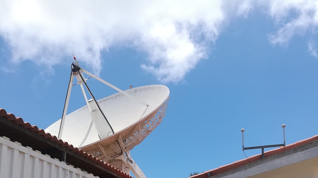 A large white satellite ground-station dish pointing at a blue sky with scattered cloud, seen over the roofs of white single-storey buildings at the Broglio Space Centre near Malindi, Kenya, on 14 February 2018; no people in view.